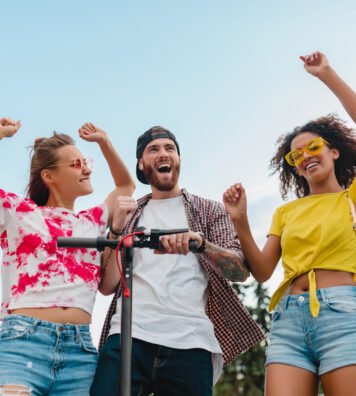 happy young company of smiling friends dancing walking in street with electric kick scooter, man and women having fun together, colorful summer hipster fashion style, talking, smiling